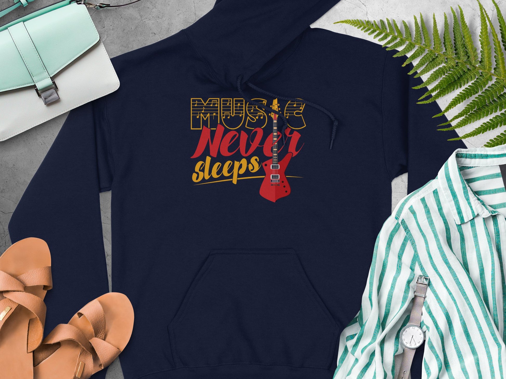 Discover comfort and style with Garment Graphics navy blue music hoodie. It features a classic fit and Music Never Sleeps alongside a guitar graphic. Pair it effortlessly with sandals, a purse, a striped shirt, wristwatch, and accentuate with fern leaves on a sleek gray surface.