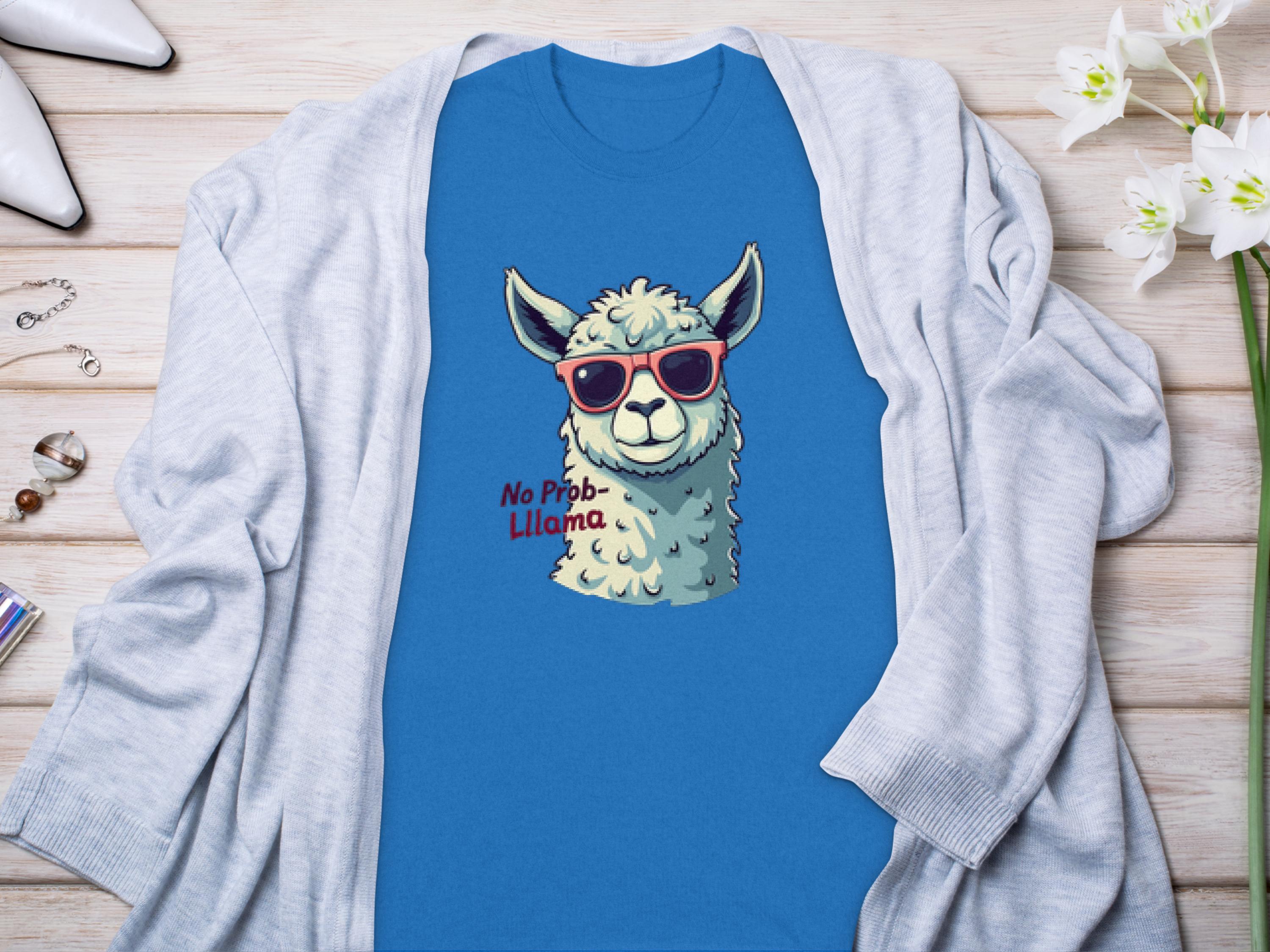 The Garment Graphics blue t-shirt flaunts a funny llama design with red sunglasses and No Prob-Llama text. Displayed on wood, its accessorized with rings, earrings, white flowers, and a light gray cardigan for charm in any wardrobe.