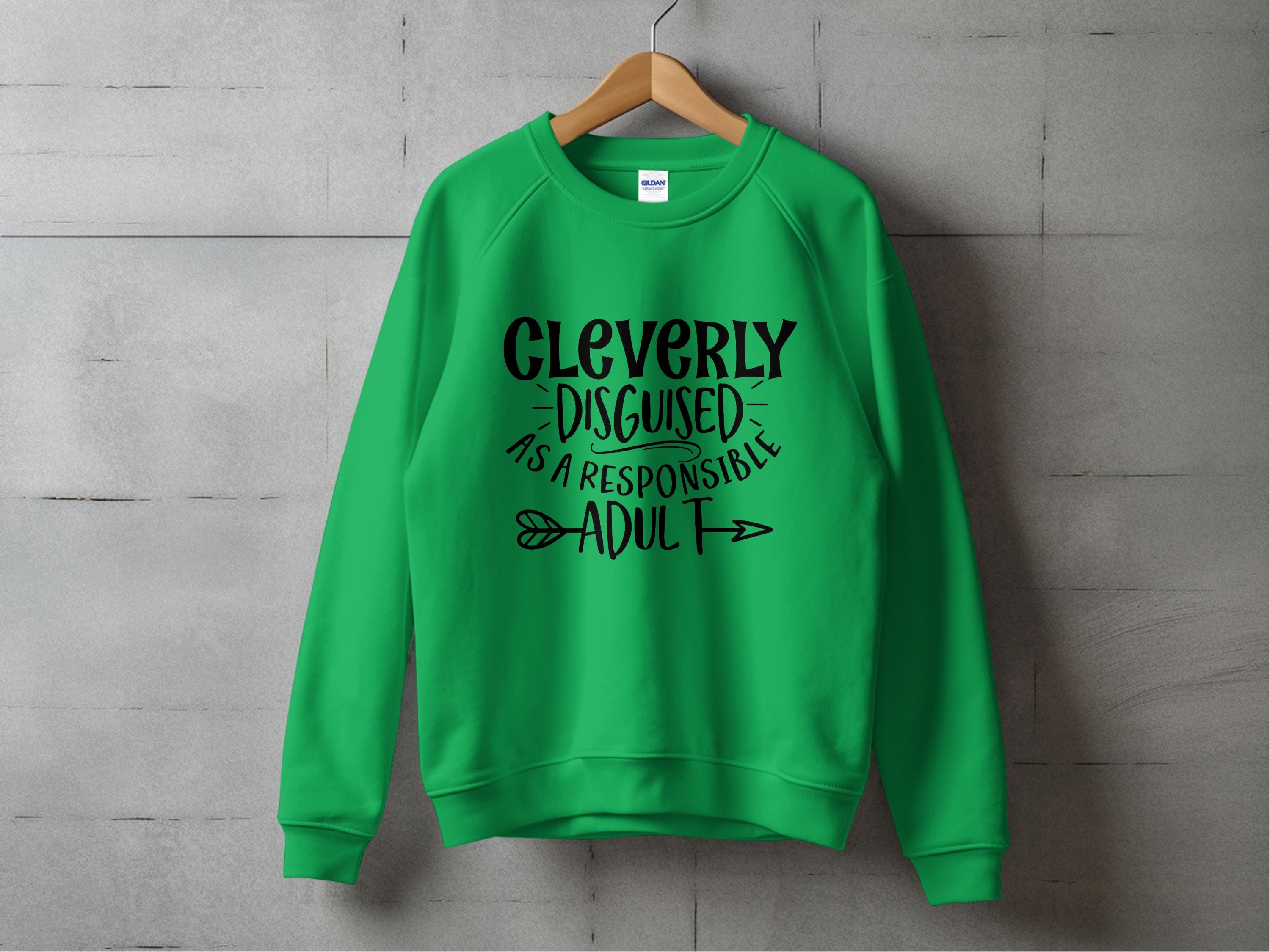 A green Garment Graphics sweatshirt displayed on a wooden hanger against a concrete wall showcases black text saying Cleverly disguised as a responsible adult with decorative arrows. This humorous piece is available in unisex sizing for anyone ready to sport it.
