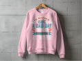 A pink fishing sweatshirt from Garment Graphics hangs on a wooden hanger against a gray textured wall, showcasing the phrase Id rather have a bad day fishing than a good day at work. Designed for fishing enthusiasts, its unisex sizing ensures comfort for everyone.