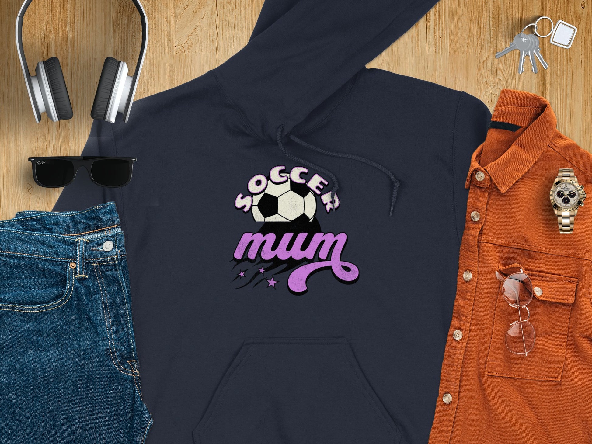 A flat lay features a Garment Graphics Soccer Mum Hoodie in classic fit with black design, paired with blue jeans, sunglasses, headphones, a wristwatch, glass cup, keys, and an orange button-up shirt on wood. Cozy and warm essentials for the sporty yet stylish routine.