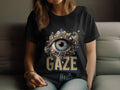 A person is sitting on a sofa wearing a black Garment Graphics T-shirt with an ornate large gemstone eye design and the bold word GAZE below. The T-shirt offers womens sizing for a perfect fit.