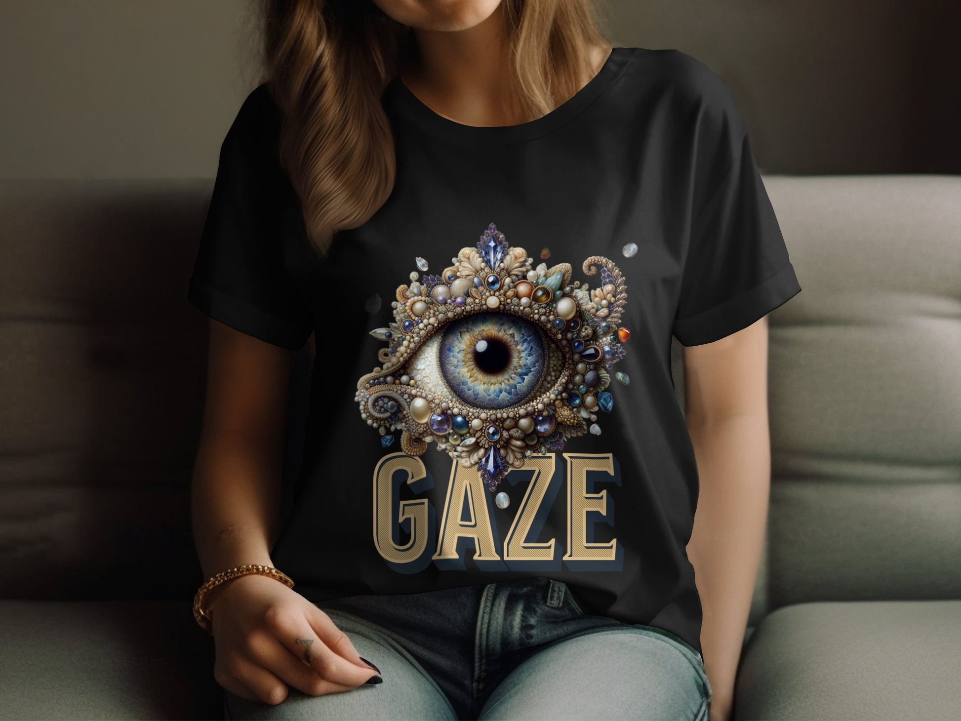 A person is sitting on a sofa wearing a black Garment Graphics T-shirt with an ornate large gemstone eye design and the bold word GAZE below. The T-shirt offers womens sizing for a perfect fit.