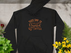 This Garment Graphics black hoodie features This Day is Powered by Coffee in brown script. Displayed flat on concrete, its beautifully framed with green plants and yellow flowers on the right and vibrant red blossoms on the left—perfect for coffee enthusiasts.
