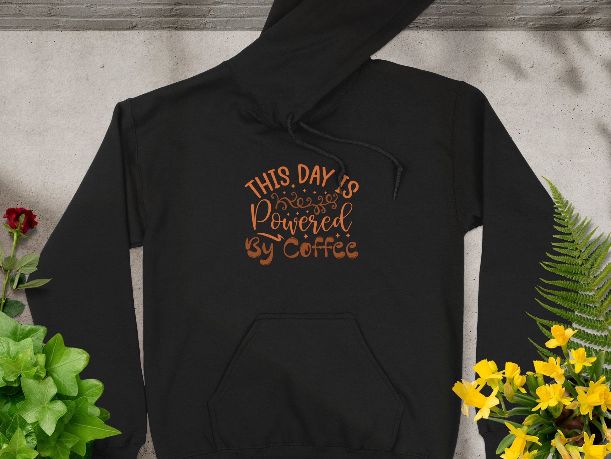 This Garment Graphics black hoodie features This Day is Powered by Coffee in brown script. Displayed flat on concrete, its beautifully framed with green plants and yellow flowers on the right and vibrant red blossoms on the left—perfect for coffee enthusiasts.