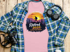 A folded pink Garment Graphics T-shirt with Retired 2025, Not My Problem Anymore and a hiking design lies under a golden sunset glow, accompanied by a blue plaid shirt, black headphones, a camera, and a coffee cup on the wooden surface.