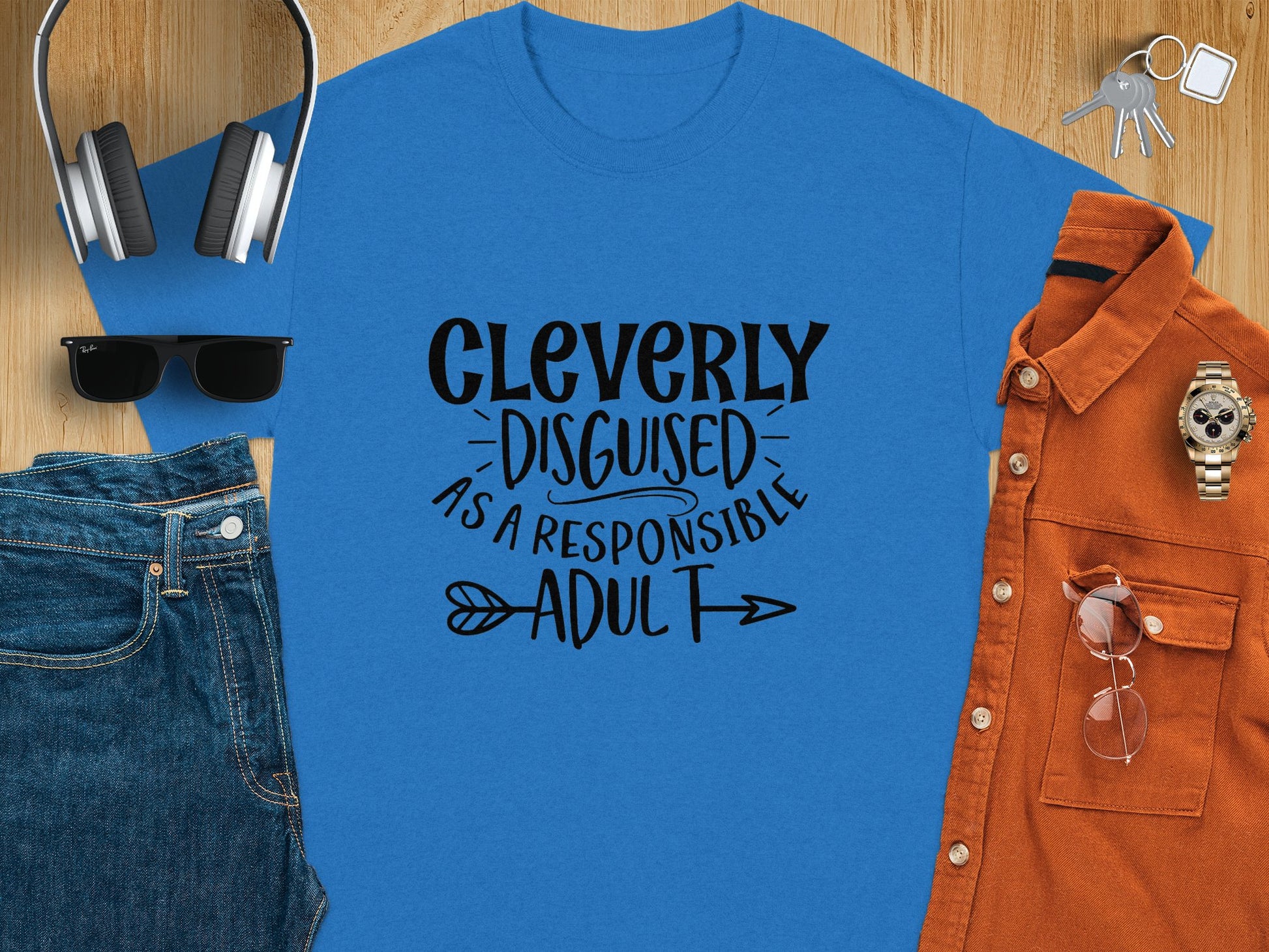 A classic fit blue t-shirt by Garment Graphics, featuring Cleverly Disguised as a Responsible Adult, is artfully displayed on wood. Ideal for casual outings, its surrounded by headphones, sunglasses, folded jeans, an orange button-up shirt, and a watch.