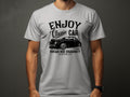 A man wears a light gray T-shirt by Garment Graphics, featuring a black classic car graphic and the text Enjoy Classic Car, Vintage Ride Community, Aircooled, Est. 1980, ideal for car enthusiasts.