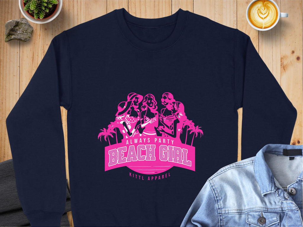 A navy blue sweatshirt by Garment Graphics showcases a pink graphic of three women among palm trees, with Always Party Beach Girl text. It promotes Kittl Apparel, and laid on wood, it pairs perfectly with a denim jacket and coffee for the ultimate Beach Girl vibe.