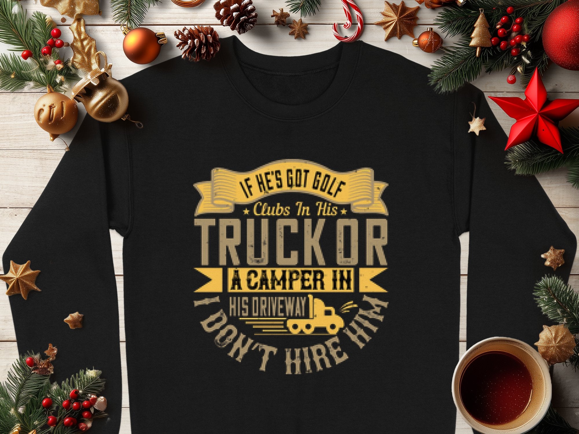 A humorous black sweatshirt by Garment Graphics, reading If hes got golf clubs in his truck or a camper in his driveway, I dont hire him, is placed on a wooden table adorned with Christmas decorations, cookies, and a cup of coffee.