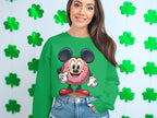 A long-haired person stands before a shamrock-adorned wall, wearing a Garment Graphics green sweatshirt featuring a colorful donut character with mouse ears. They smile with hands in pockets, highlighting the sweatshirts comfortable unisex sizing.