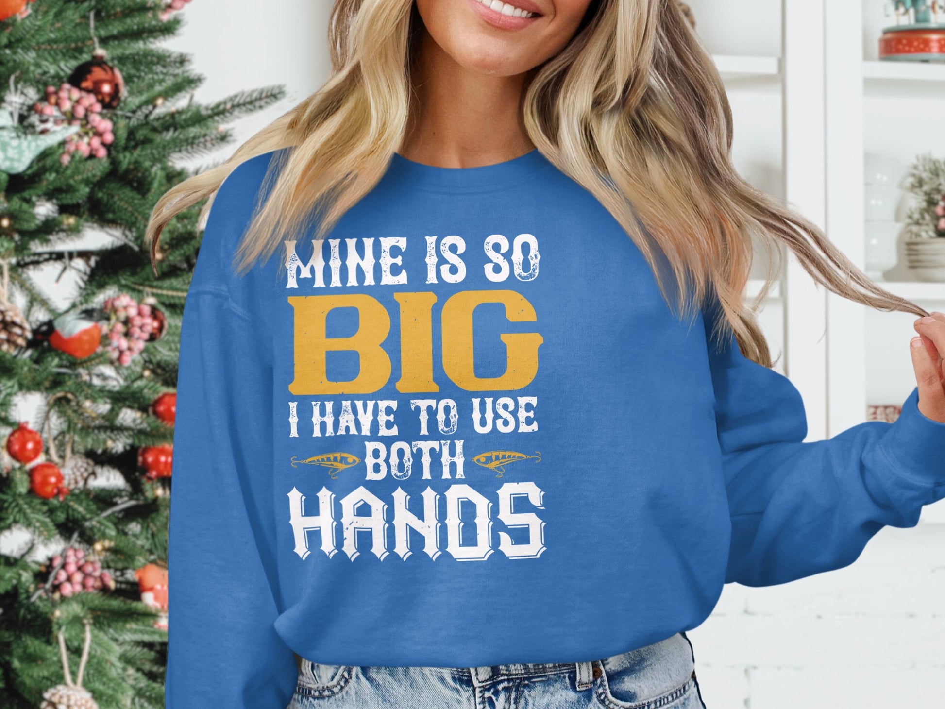 A person poses by a Christmas tree, wearing a blue sweatshirt from Garment Graphics with the bold statement MINE IS SO BIG I HAVE TO USE BOTH HANDS in white and gold letters displayed prominently, casually holding a strand of hair.