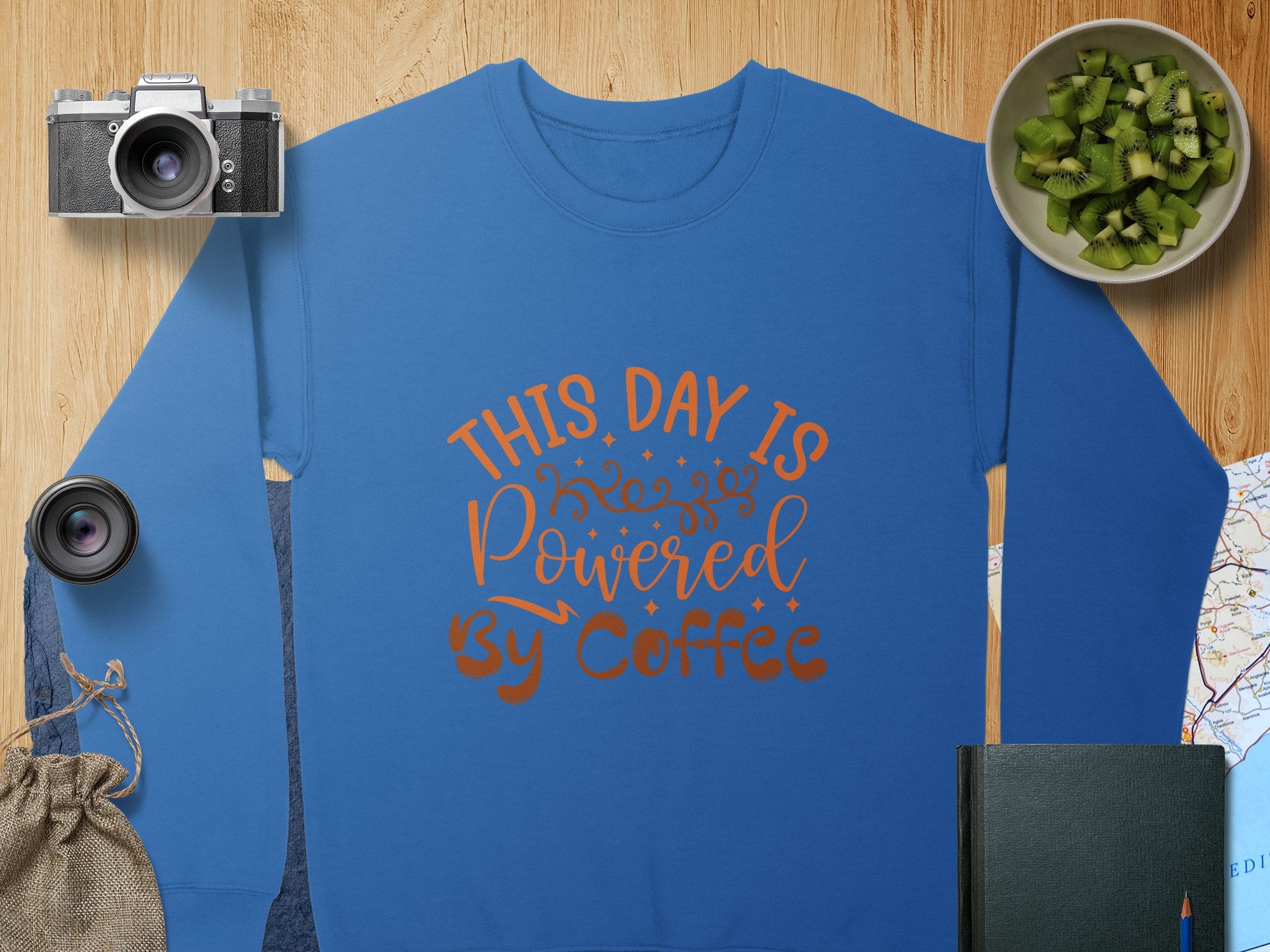 A Garment Graphics blue graphic sweatshirt with This Day is Powered by Coffee in orange lettering sits on a wooden surface. Nearby are a camera, lens, kiwi bowl, map, notebook, and drawstring pouch creating an inviting scene.