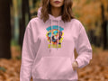 A person wears a vibrant light pink hoodie by Garment Graphics, featuring a dog with sunglasses and the text Pawsitive Stylish, set against a blurred backdrop of orange fall leaves and tree trunks, creating a unique style.