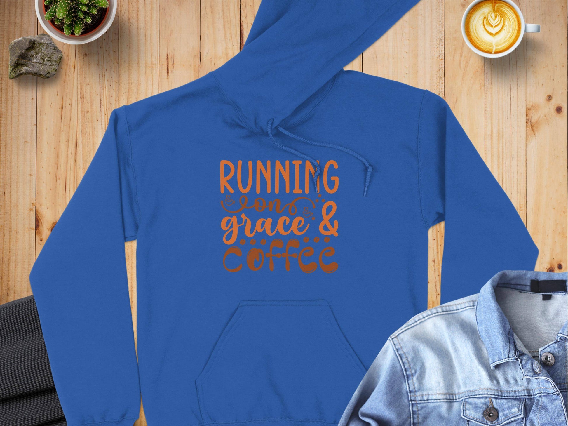 A blue Garment Graphics hoodie with Running on grace & coffee in orange lettering lies on a wooden surface. Its crafted from medium-heavy fabric with a classic fit. Nearby are a latte art coffee cup, denim jacket, and small potted plant.