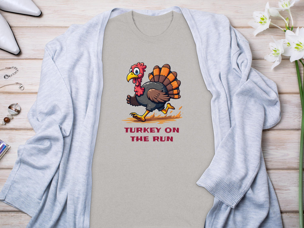 A Garment Graphics T-shirt with a cartoon turkey and Turkey on the Run print, styled with a light gray cardigan, displayed alongside accessories and white flowers on wood. Available in family sizes.