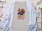 A Garment Graphics T-shirt with a cartoon turkey and Turkey on the Run print, styled with a light gray cardigan, displayed alongside accessories and white flowers on wood. Available in family sizes.