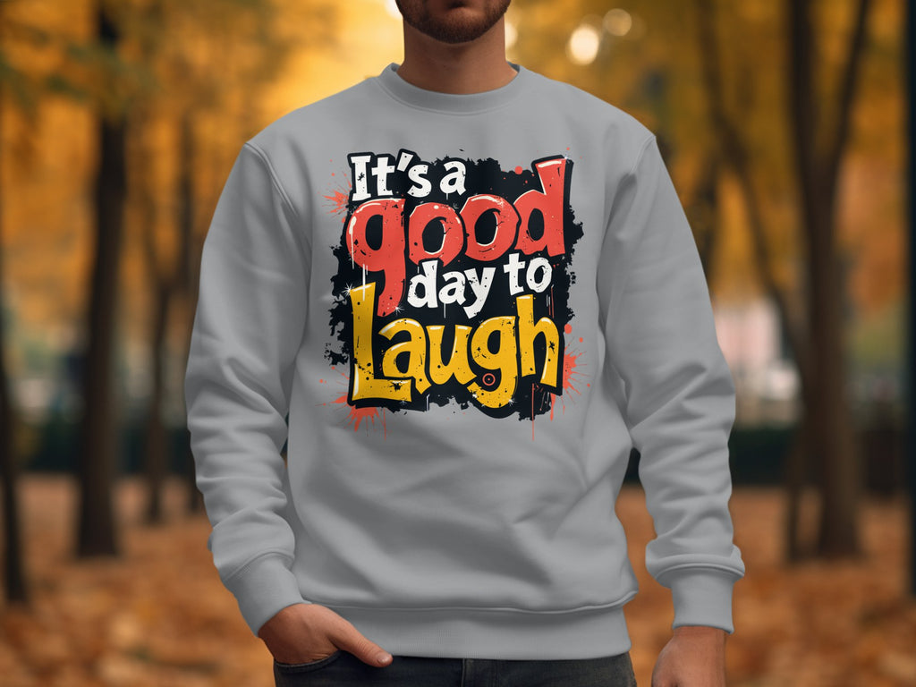 A person in a light gray Garment Graphics sweatshirt, featuring the vibrant text Its a Good Day to Laugh, stands in a park amid autumn leaves, with fall foliage creating a picturesque backdrop.