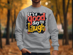 A person in a light gray Garment Graphics sweatshirt, featuring the vibrant text Its a Good Day to Laugh, stands in a park amid autumn leaves, with fall foliage creating a picturesque backdrop.