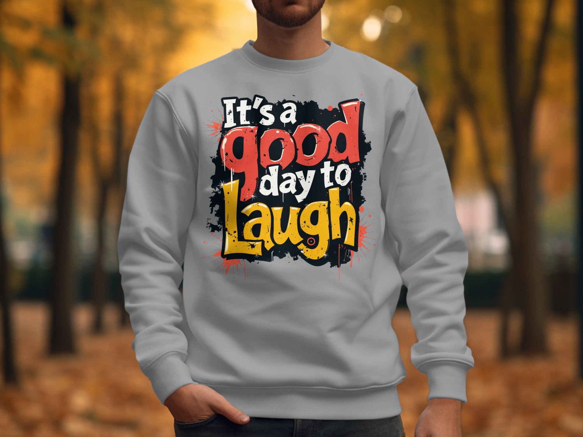 A person in a light gray Garment Graphics sweatshirt, featuring the vibrant text Its a Good Day to Laugh, stands in a park amid autumn leaves, with fall foliage creating a picturesque backdrop.
