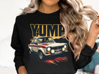 A person with long, wavy hair dons a Garment Graphics black sweatshirt with a rally car and bold YUMP print. They hold a strand of hair, embracing the classic designs appeal.