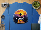 The Garment Graphics retirement sweatshirt is blue and features a hiker design with a sunset, trees, and mountains silhouette. The text Retired 2025: Not My Problem Anymore is surrounded by illustrations of a camera, lens, cucumbers, map, book, and pouch. Available in unisex sizing.