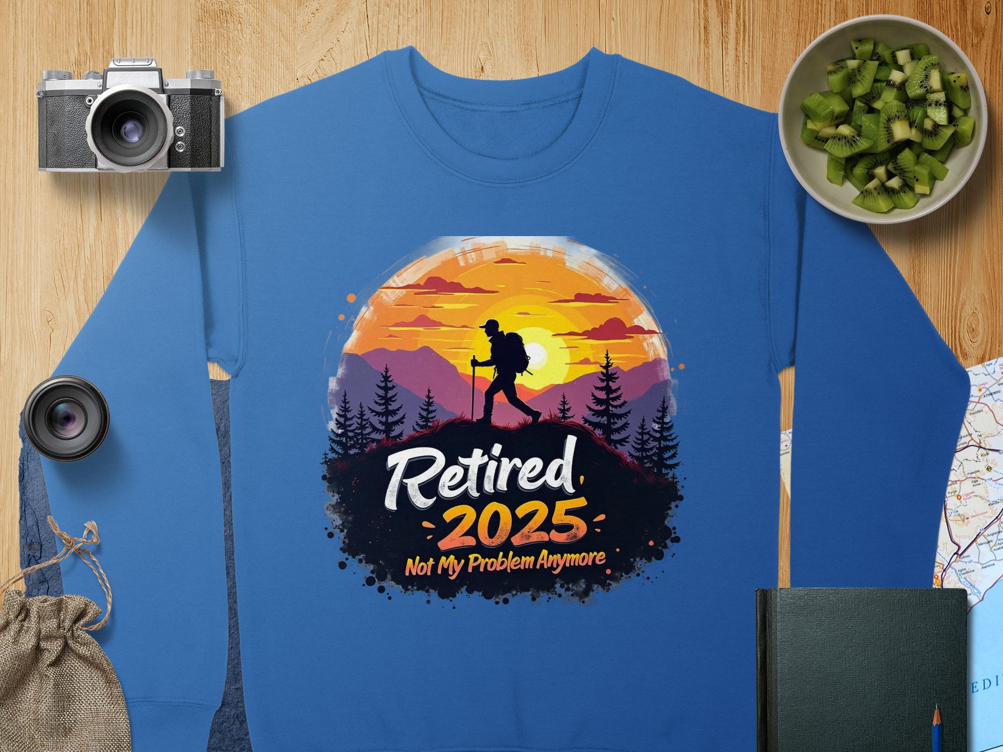 The Garment Graphics retirement sweatshirt is blue and features a hiker design with a sunset, trees, and mountains silhouette. The text Retired 2025: Not My Problem Anymore is surrounded by illustrations of a camera, lens, cucumbers, map, book, and pouch. Available in unisex sizing.