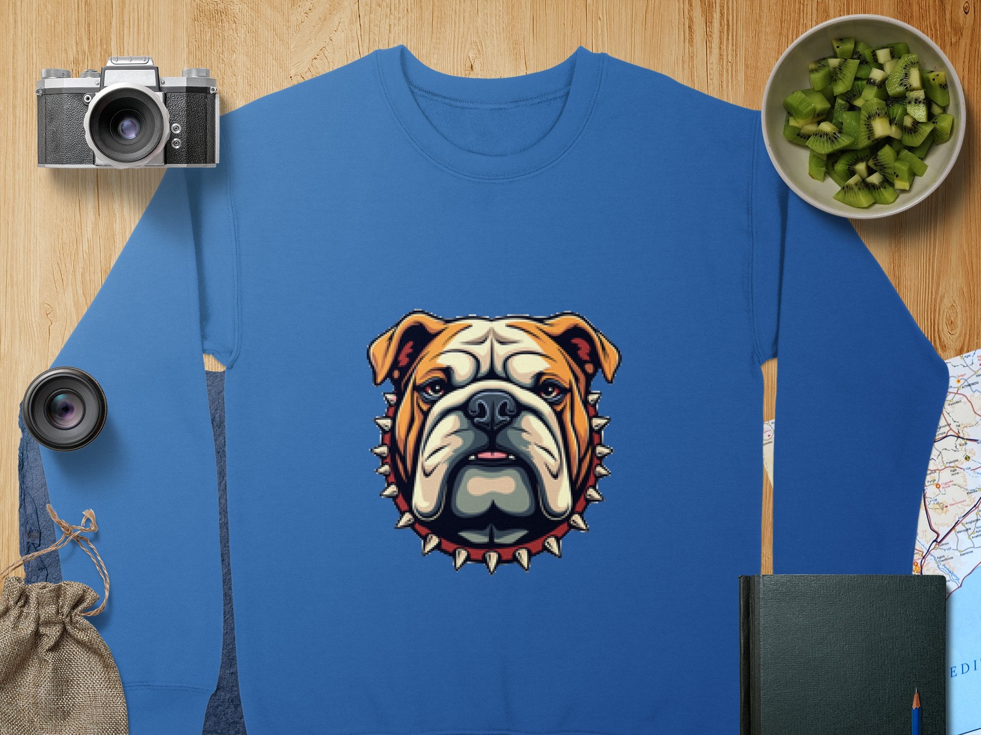 The Garment Graphics blue sweatshirt boasts a bold bulldog graphic with a spiked collar and is perfect for relaxed unisex wear. Its styled alongside items like a camera, lens, kiwi bowl, jute bag, book, and map on wood.