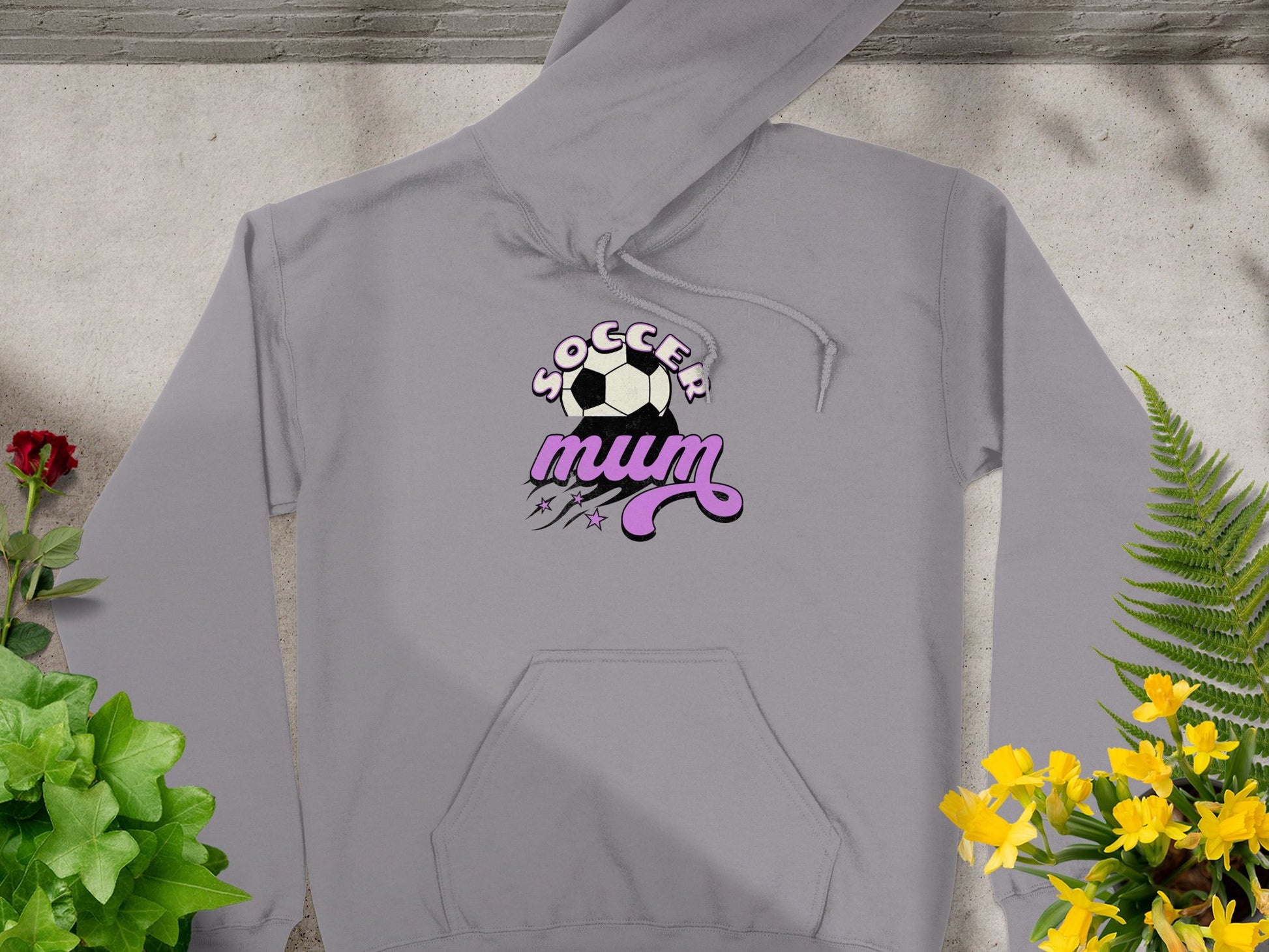 A cozy gray hoodie from Garment Graphics, featuring a soccer ball and Soccer Mum in white and purple, lies flat against green foliage with vibrant yellow and red flowers, offering a classic fit.