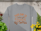 A Garment Graphics gray T-shirt features the fun design This Day is Powered by Coffee in orange cursive. Its displayed on a concrete surface with surrounding green, red, and yellow plants, while shadows play across the scene.