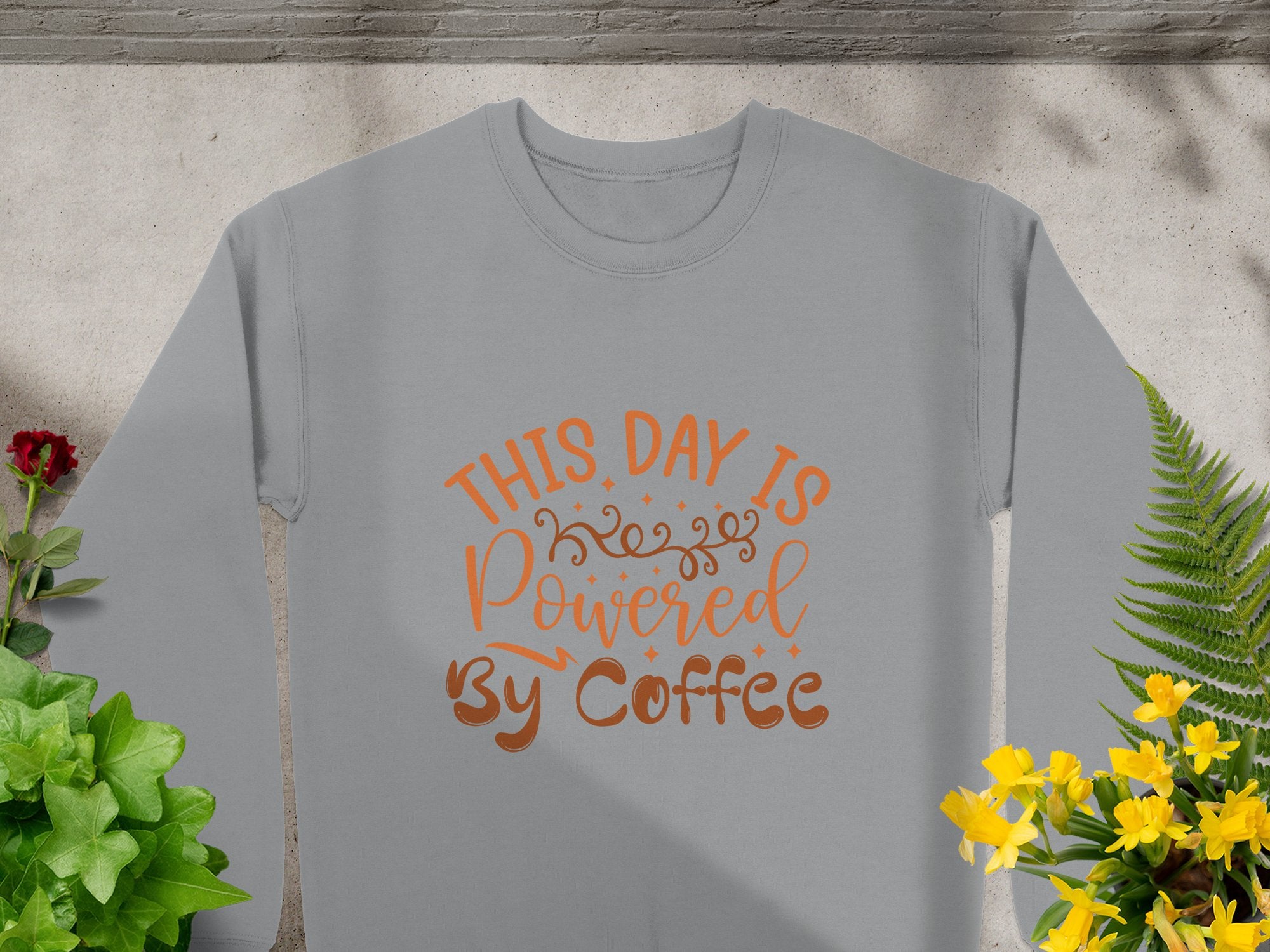 A Garment Graphics gray T-shirt features the fun design This Day is Powered by Coffee in orange cursive. Its displayed on a concrete surface with surrounding green, red, and yellow plants, while shadows play across the scene.