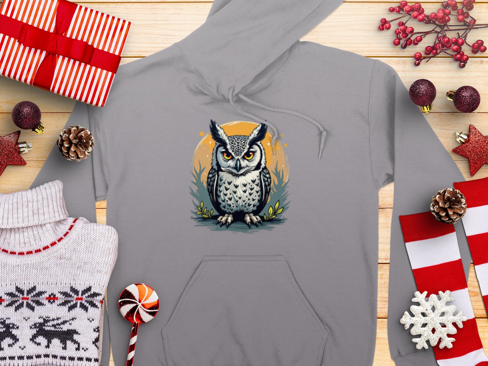 A Garment Graphics gray hoodie with a fierce cartoon owl is displayed with festive decorations like gifts, pinecones, and candy canes. A cozy scene in the lower left features a folded sweater with deer patterns made from medium-heavy fabric.