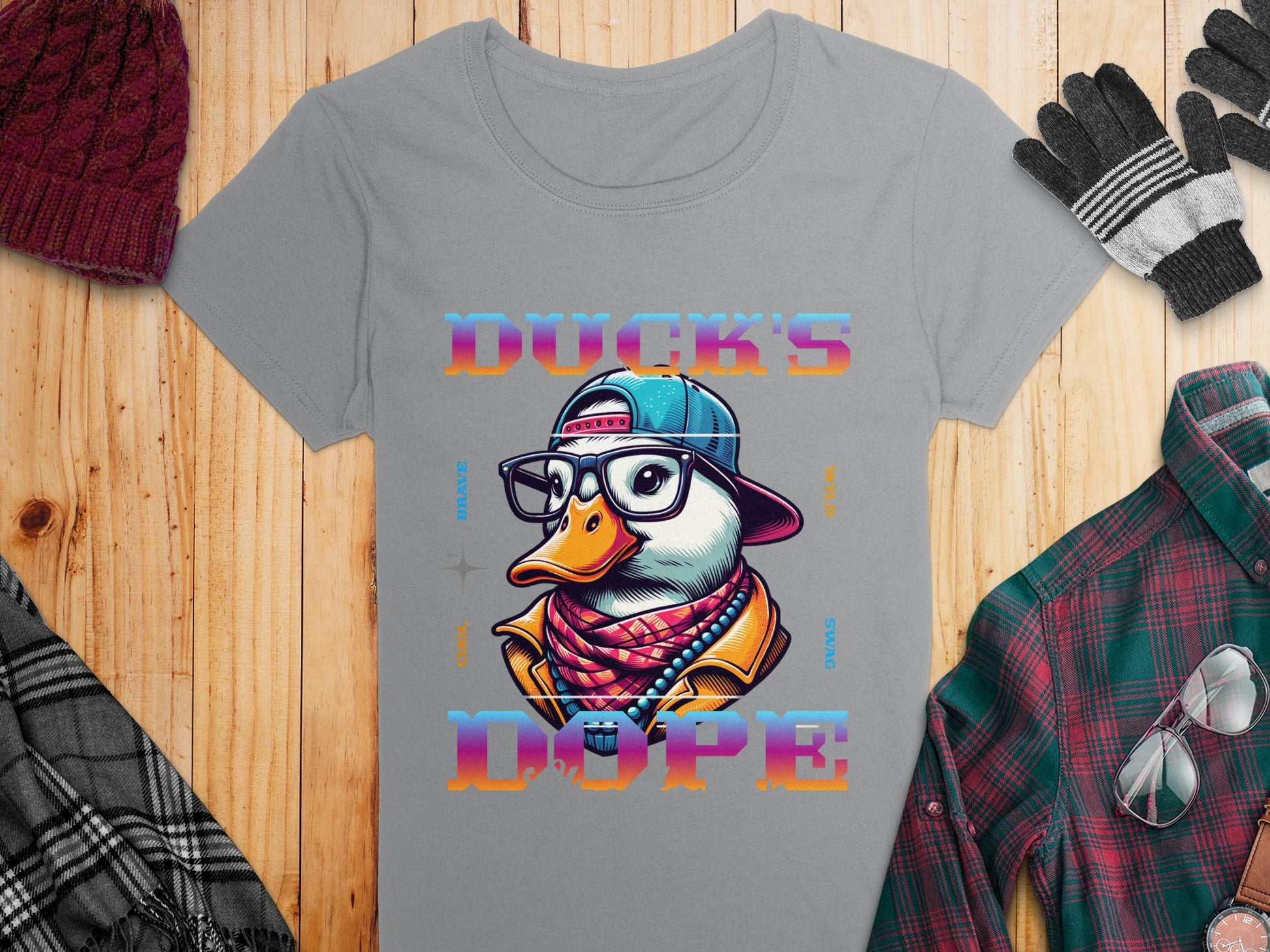 A gray T-shirt from Garment Graphics features a cartoon duck with a cap, glasses, and scarf on a wooden surface. The colorful text Ducks Dope enhances its design. It sits among plaid shirts, gloves, and a knitted hat.