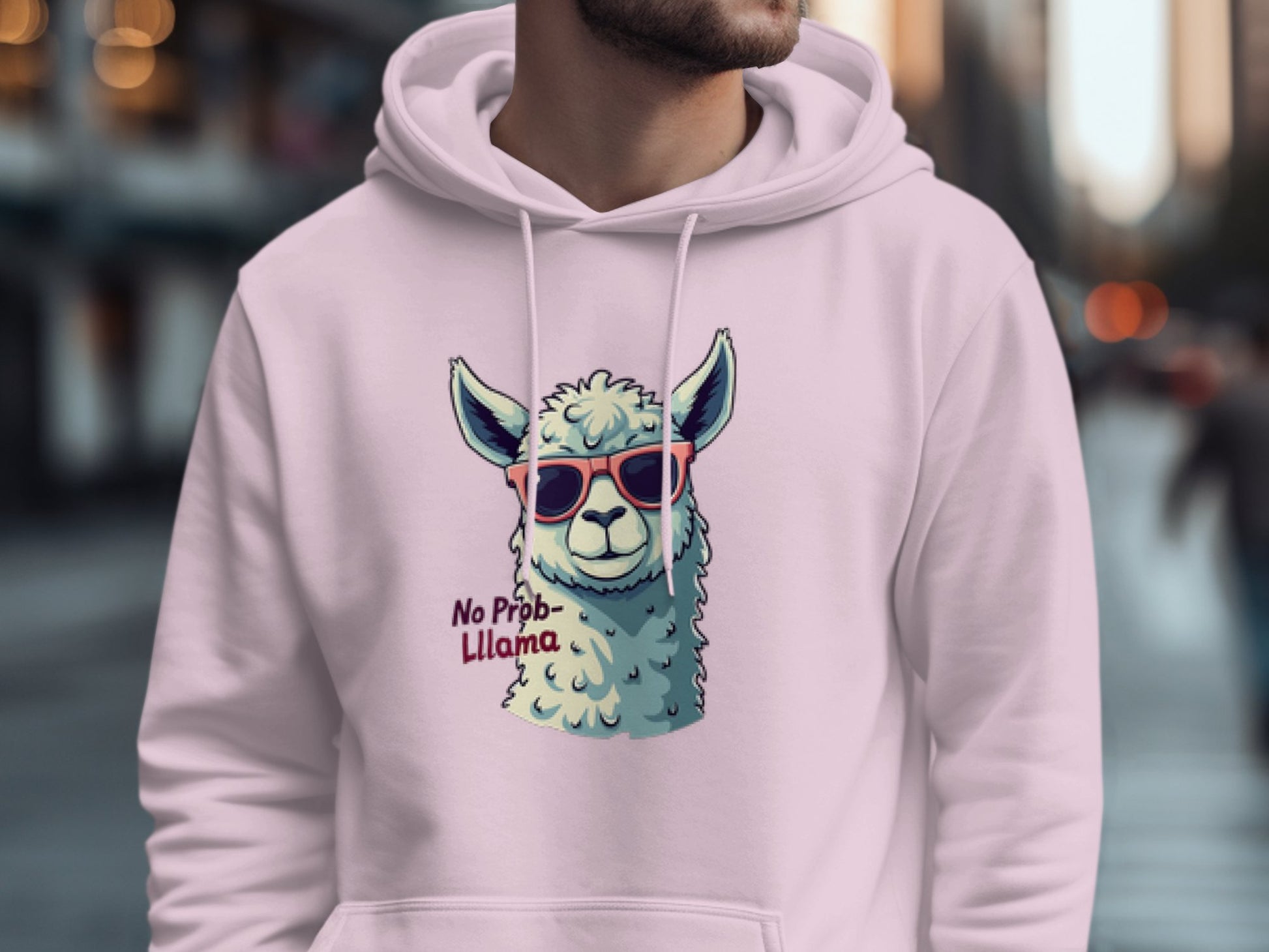 A person wears a light pink graphic hoodie from Garment Graphics, featuring a llama in red sunglasses and the slogan No Prob-Llama, making it pop against the blurred urban street backdrop.