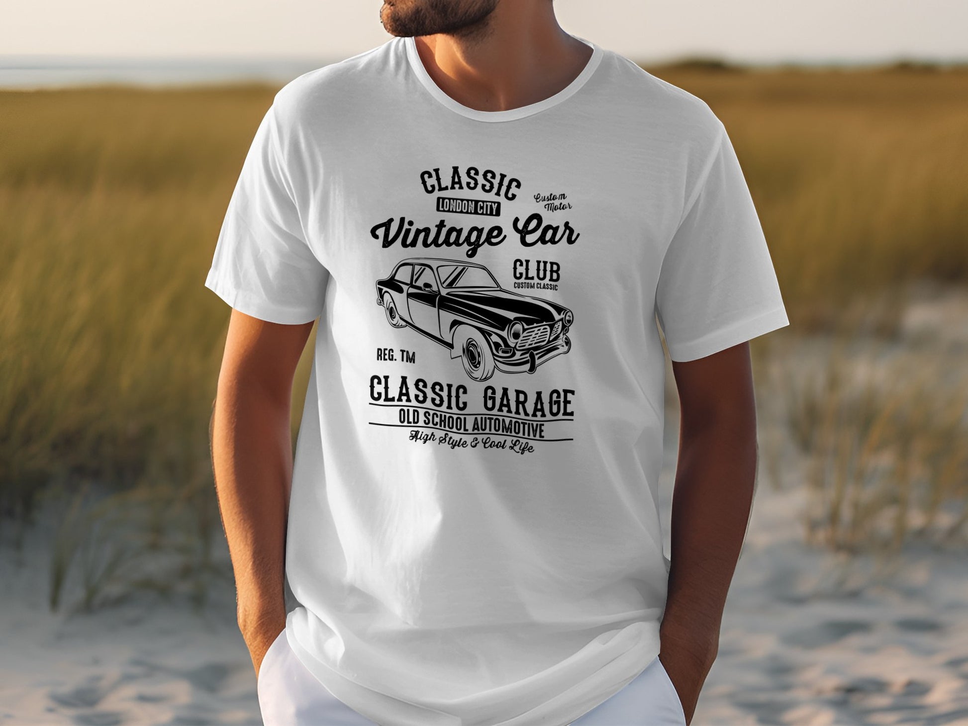Someone is wearing a Garment Graphics classic fit white T-shirt, featuring a graphic of a vintage car with text: Classic London City Vintage Car Club, Classic Garage, and Old School Automotive. A grassy field serves as the backdrop.