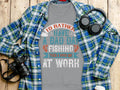 A gray Id Rather Have a Bad Day Fishing Than a Good Day at Work T-shirt from Garment Graphics rests on a wooden surface, surrounded by a blue plaid shirt, headphones, and a camera.