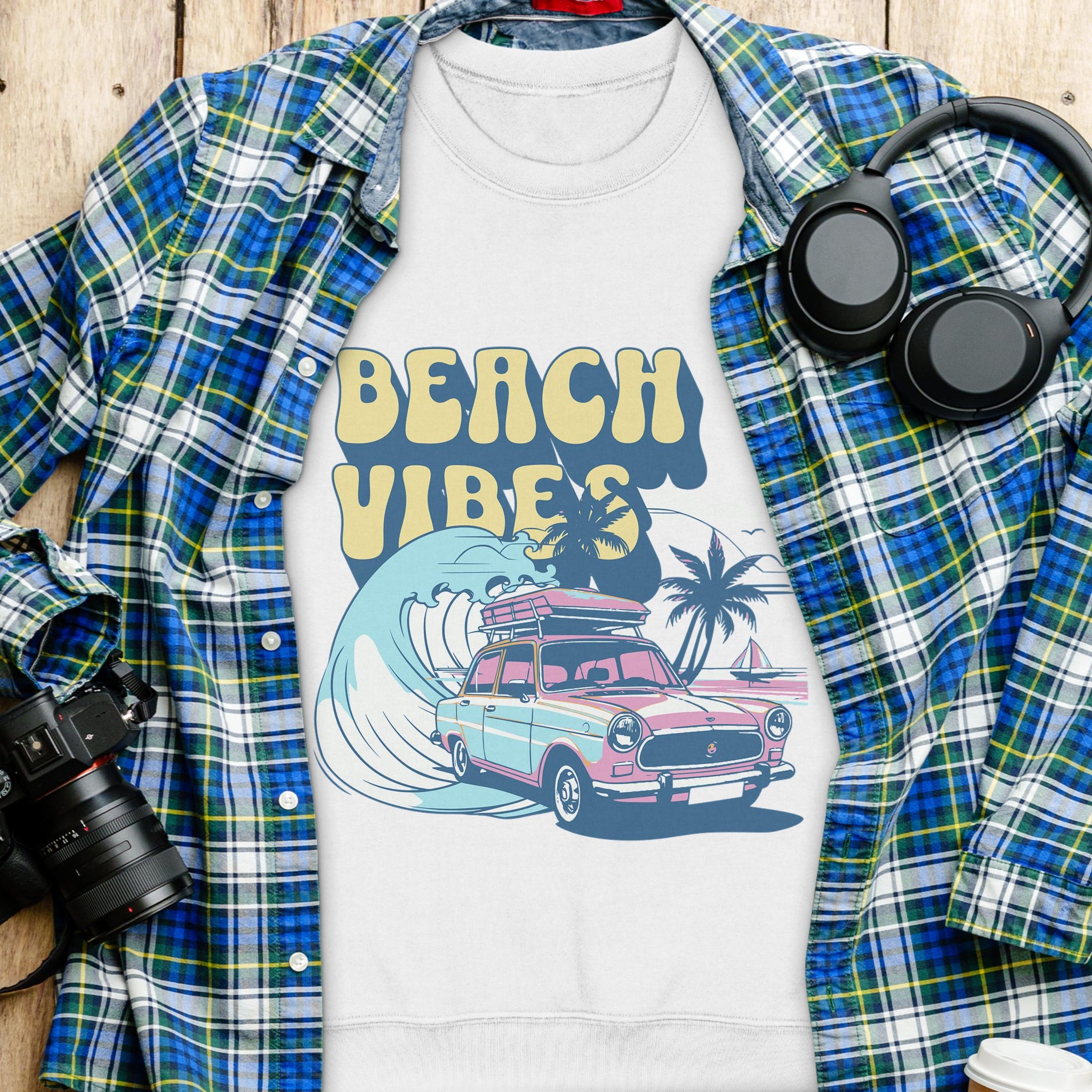 A white Garment Graphics T-shirt featuring a retro car, surfboards, palm trees, and Beach Vibes text in unisex sizing is displayed on a wooden surface with a plaid shirt, headphones, camera, and white cup, capturing the essence of a retro road trip.