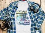 A white Garment Graphics T-shirt featuring a retro car, surfboards, palm trees, and Beach Vibes text in unisex sizing is displayed on a wooden surface with a plaid shirt, headphones, camera, and white cup, capturing the essence of a retro road trip.