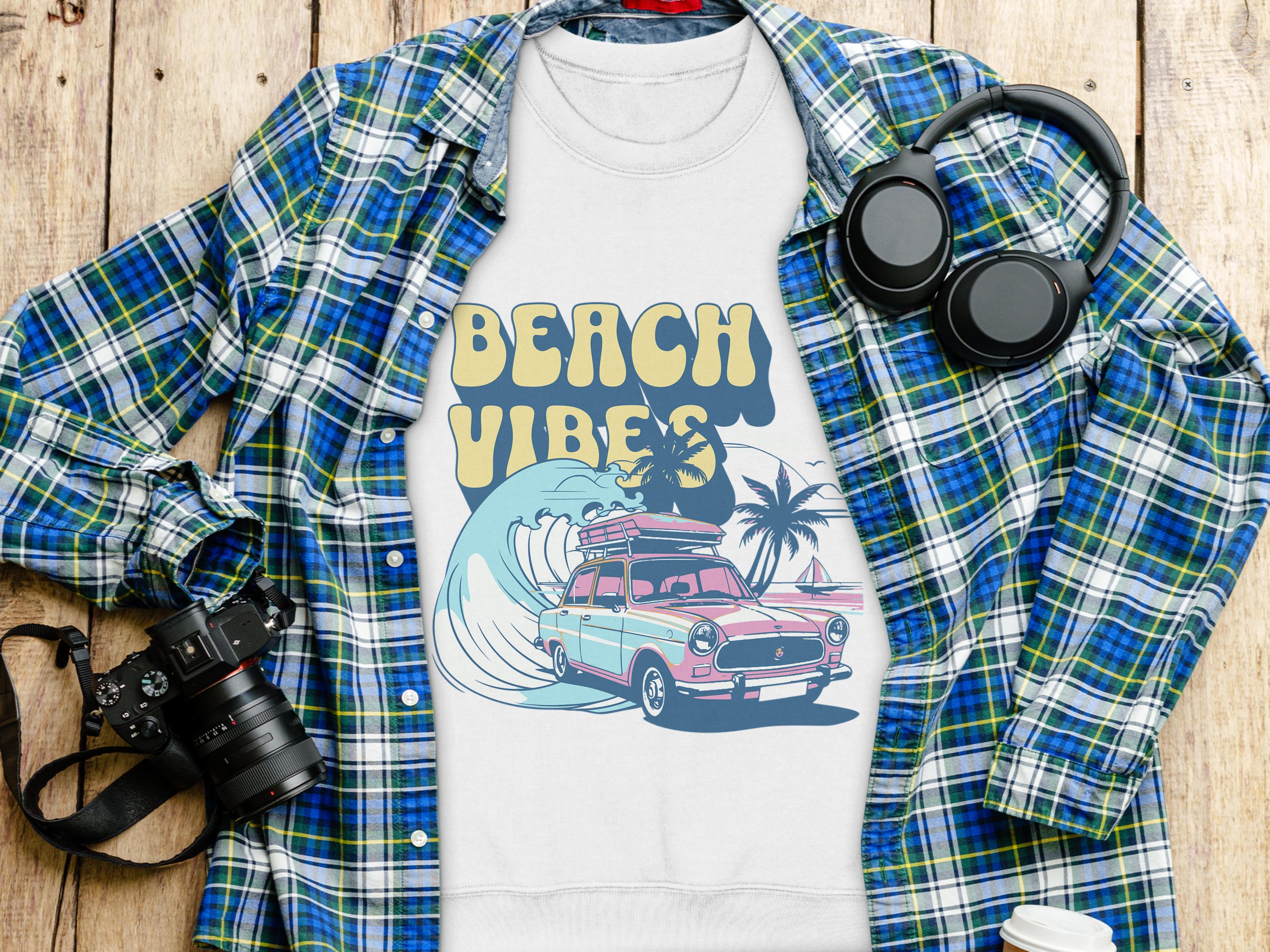 A white Garment Graphics T-shirt featuring a retro car, surfboards, palm trees, and Beach Vibes text in unisex sizing is displayed on a wooden surface with a plaid shirt, headphones, camera, and white cup, capturing the essence of a retro road trip.