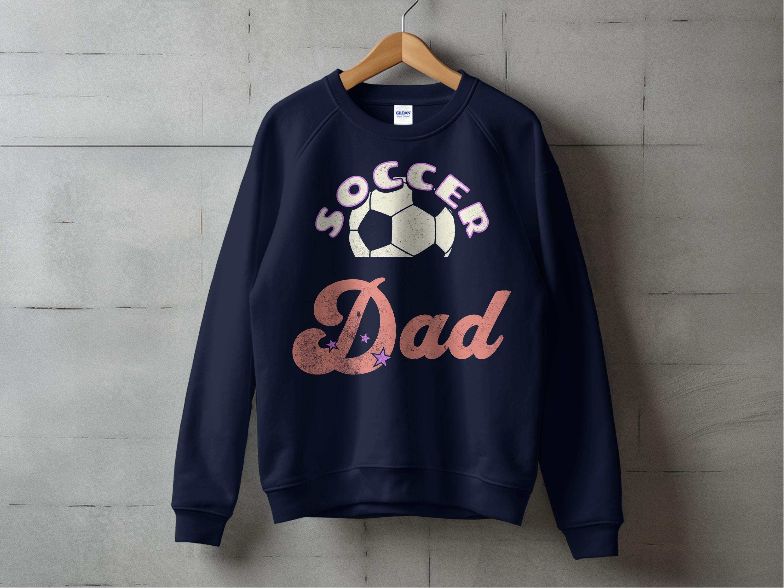 A stylish unisex navy Soccer Dad sweatshirt from Garment Graphics hangs on a wooden hanger against a concrete wall, featuring a bold stylized Soccer Dad text and a soccer ball graphic.