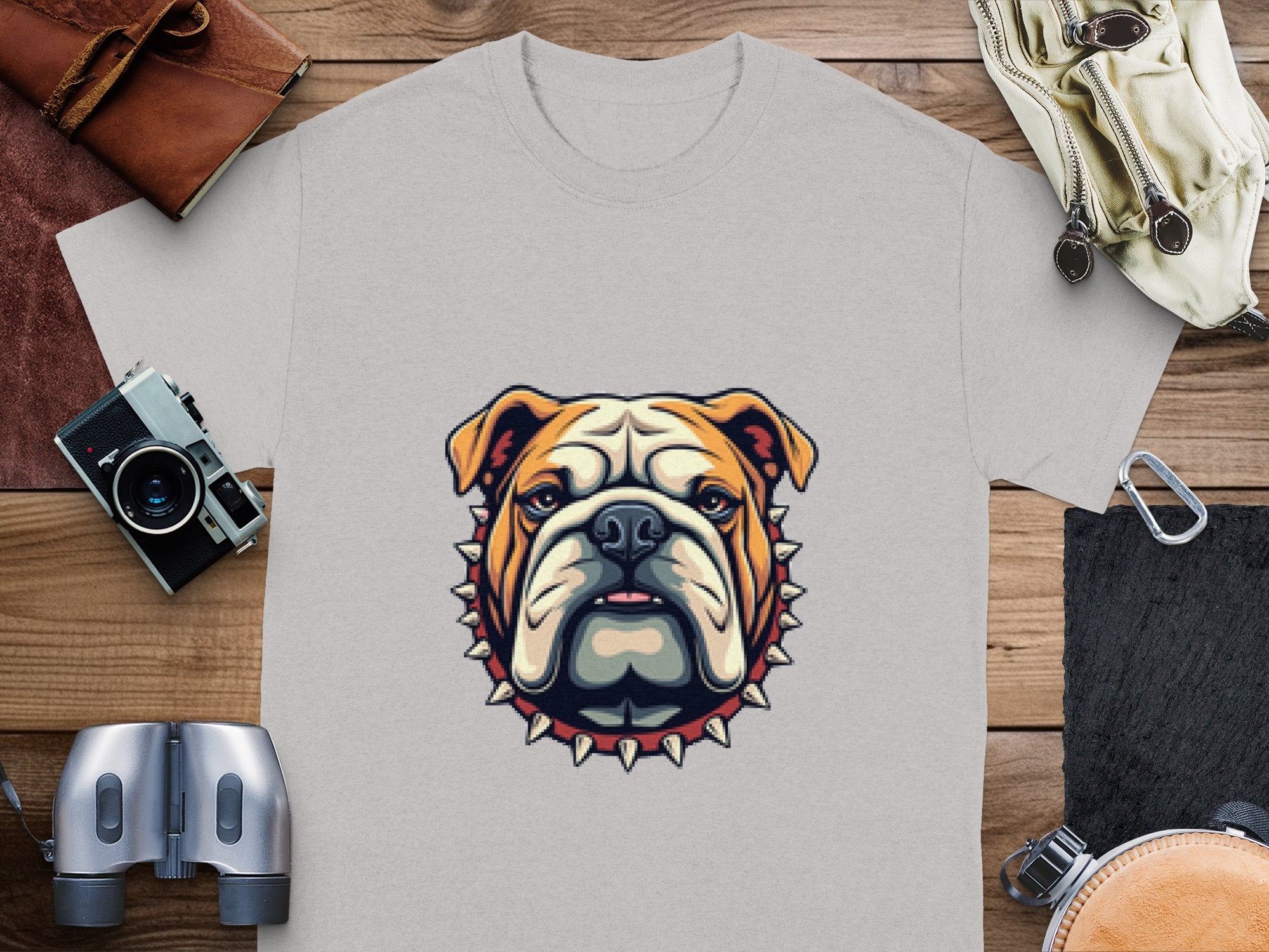 A bulldog graphic T-shirt in cool gray from Garment Graphics, with a spiked collar design, sits on a wooden surface. Its surrounded by a vintage camera, leather wallets, binoculars, and gloves—ideal for fans of unique style and available in family sizes.