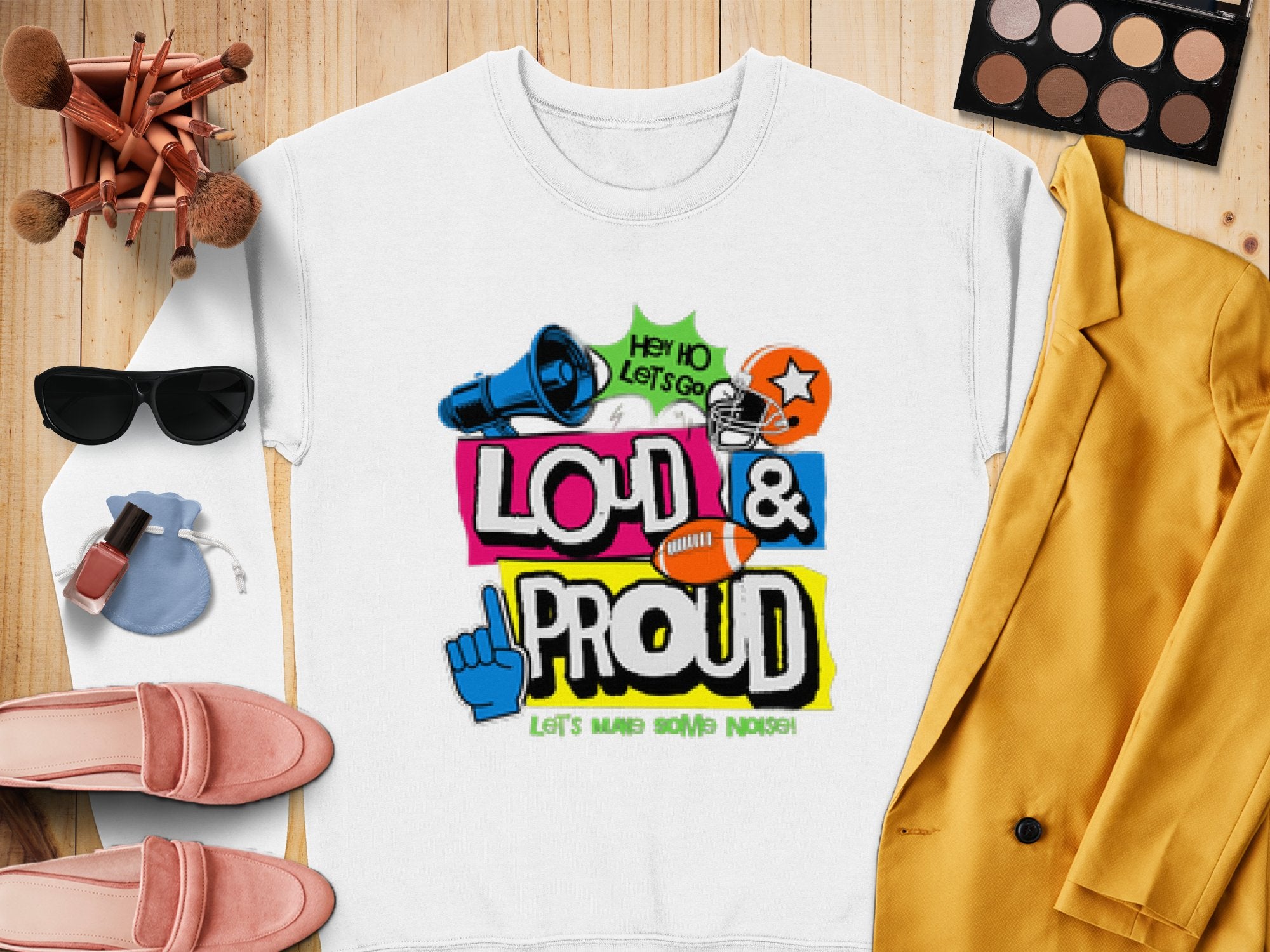 A white tee by Garment Graphics features Loud & Proud text in color, with blue fist, megaphone, star, and sports icons. Resting on wood beside a yellow jacket, pink shoes, and shades, this unique style matches the unisex fit of Loud and Proud sweatshirts seamlessly.