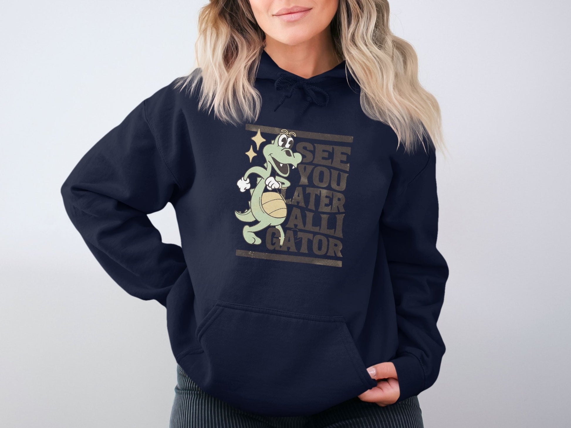 A person wearing a classic fit, navy blue See You Later Alligator hoodie by Garment Graphics, featuring a playful gator graphic, stands against a plain white background. Their wavy, light-colored hair complements the casual vibe of this fun attire.