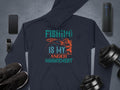 A dark blue hoodie from Garment Graphics Anglers Wardrobe collection, with Fishing is my anger management in green, orange, and white, is displayed with a smartphone, smartwatch, black sneakers, dumbbells, and a portable speaker on a gray background.