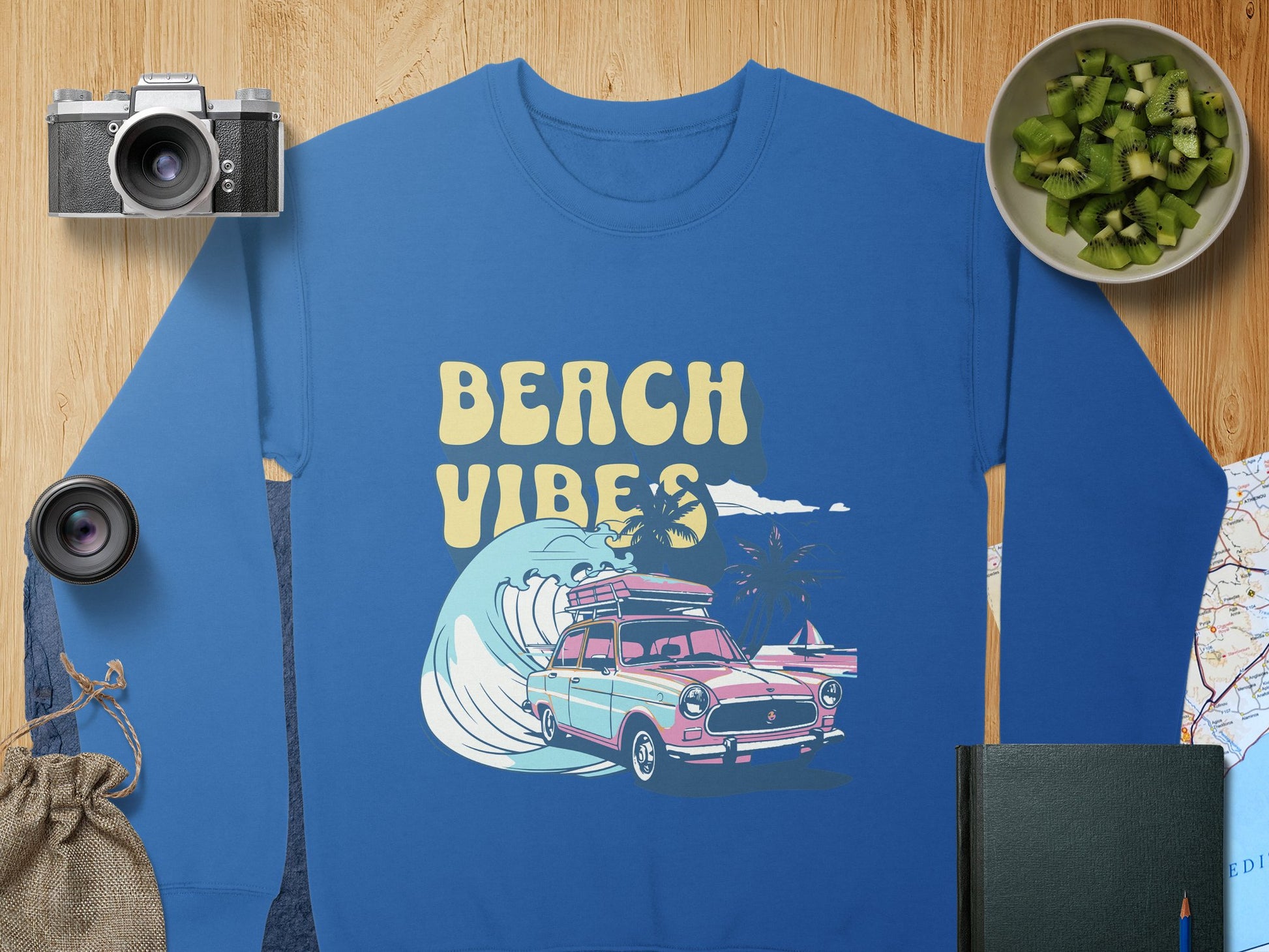 A Garment Graphics blue sweatshirt featuring a Beach Vibes design with a classic car, surfboards, and ocean waves. Nearby are a camera, lens, kiwi in a green bowl, notebook, map, and burlap sack on wood. Ideal for retro seaside vibes.