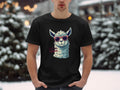 In a snowy outdoor scene, a man wears a black Garment Graphics T-shirt from the Llama T-Shirts collection, showcasing an illustrated llama with sunglasses and the text No Prob-Llama. His hands rest in his pockets, and snow-covered trees are in view behind him.