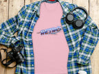 A pink Garment Graphics T-shirt with RETRO in blue and red text exuding vintage charm is displayed on a wooden surface, accompanied by a blue and green plaid shirt, camera, headphones, and a coffee cup lid.