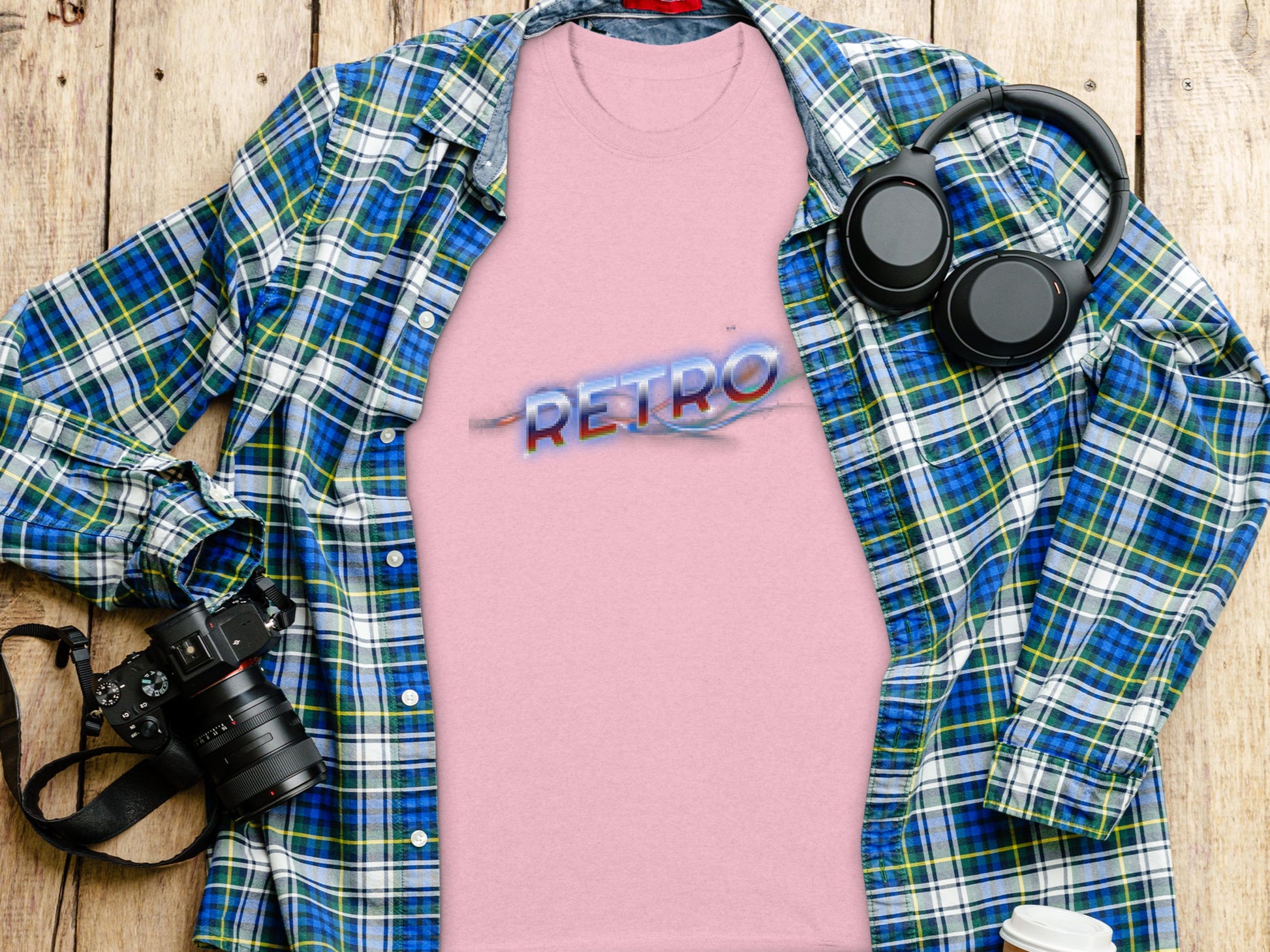 A pink Garment Graphics T-shirt with RETRO in blue and red text exuding vintage charm is displayed on a wooden surface, accompanied by a blue and green plaid shirt, camera, headphones, and a coffee cup lid.