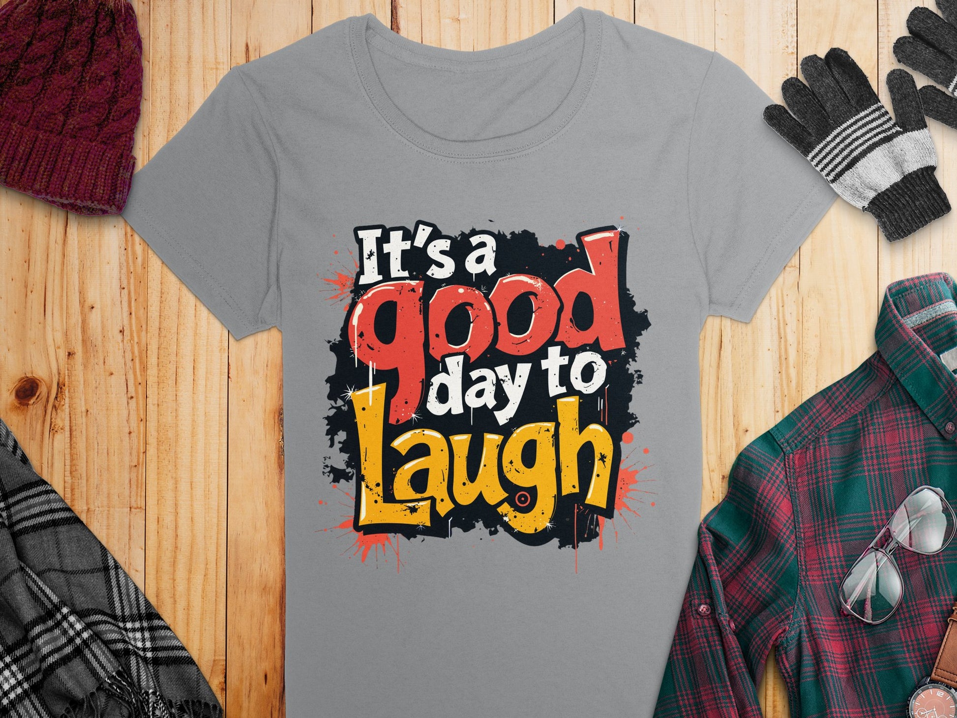 Garment Graphics offers a semi-fitted gray t-shirt with a vibrant Its a good day to Laugh graphic. Displayed with a knit hat, gloves, scarf, and plaid shirt on wooden backdrop, it exudes positivity.