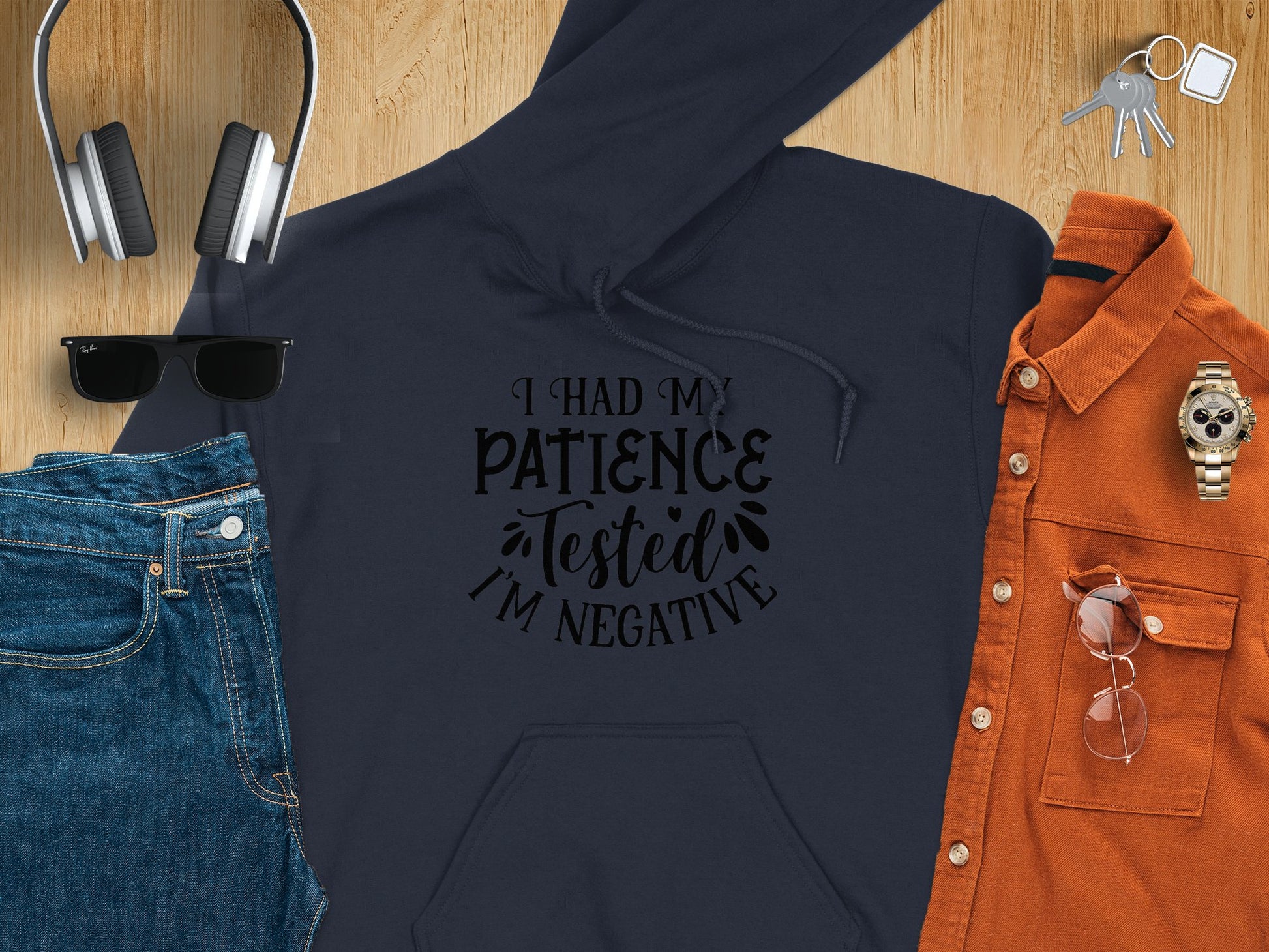 The Garment Graphics black novelty graphic hoodie showcases I had my patience tested Im negative in a decorative font. It complements jeans, sunglasses, headphones, a watch, a red-orange button-up shirt, and keys on a wood surface—a perfect blend of unique design and style.